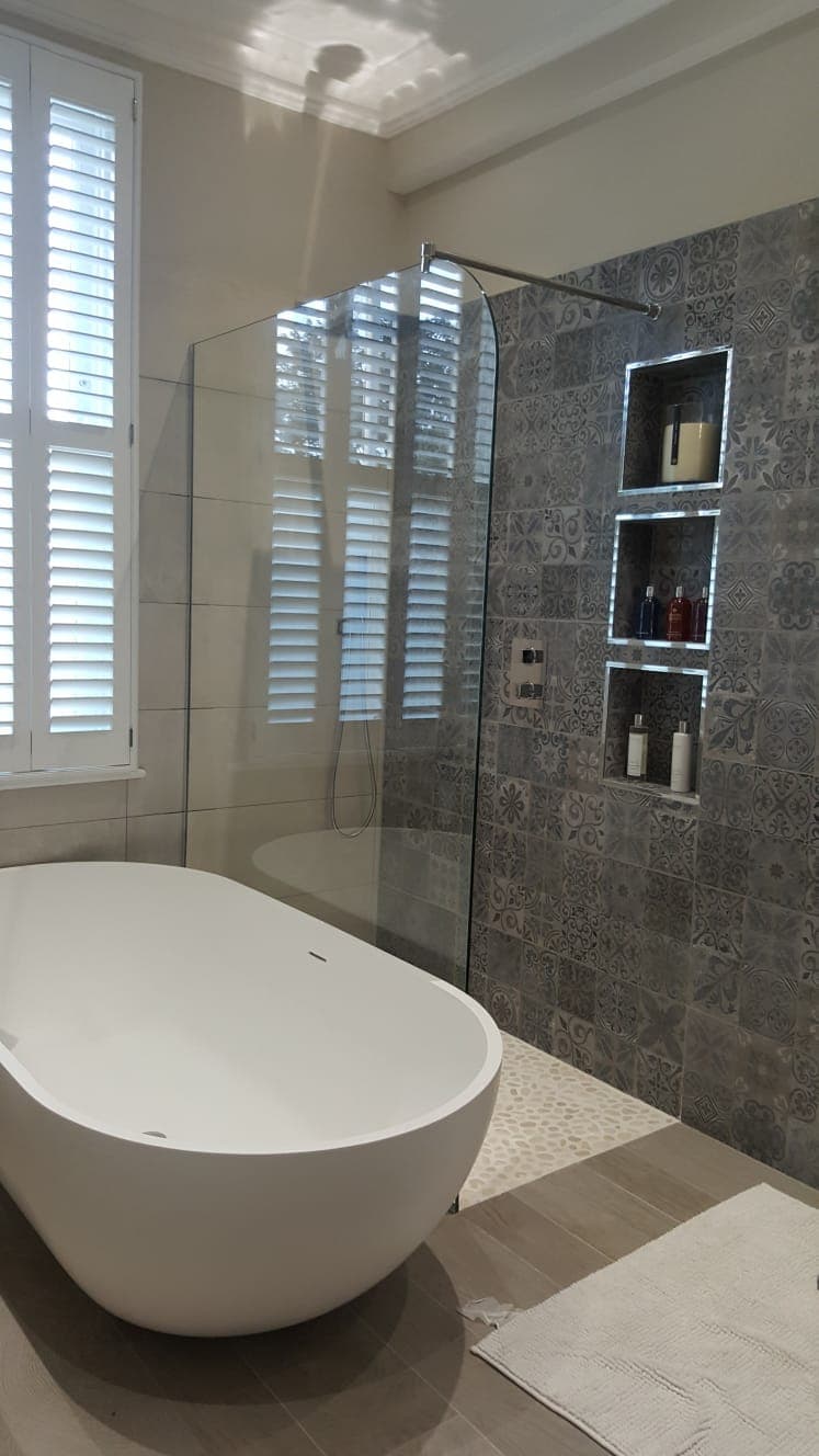 Walk-in shower screen next to a freestanding oval bath, with patterned grey wall tiles and recessed shelving niches