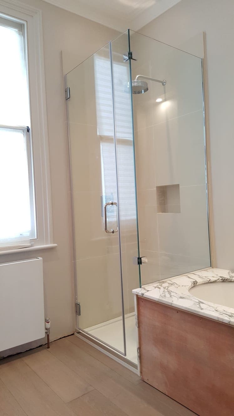 Hinged frameless shower door with a long ladder handle, beside a stone-topped vanity and a sash window