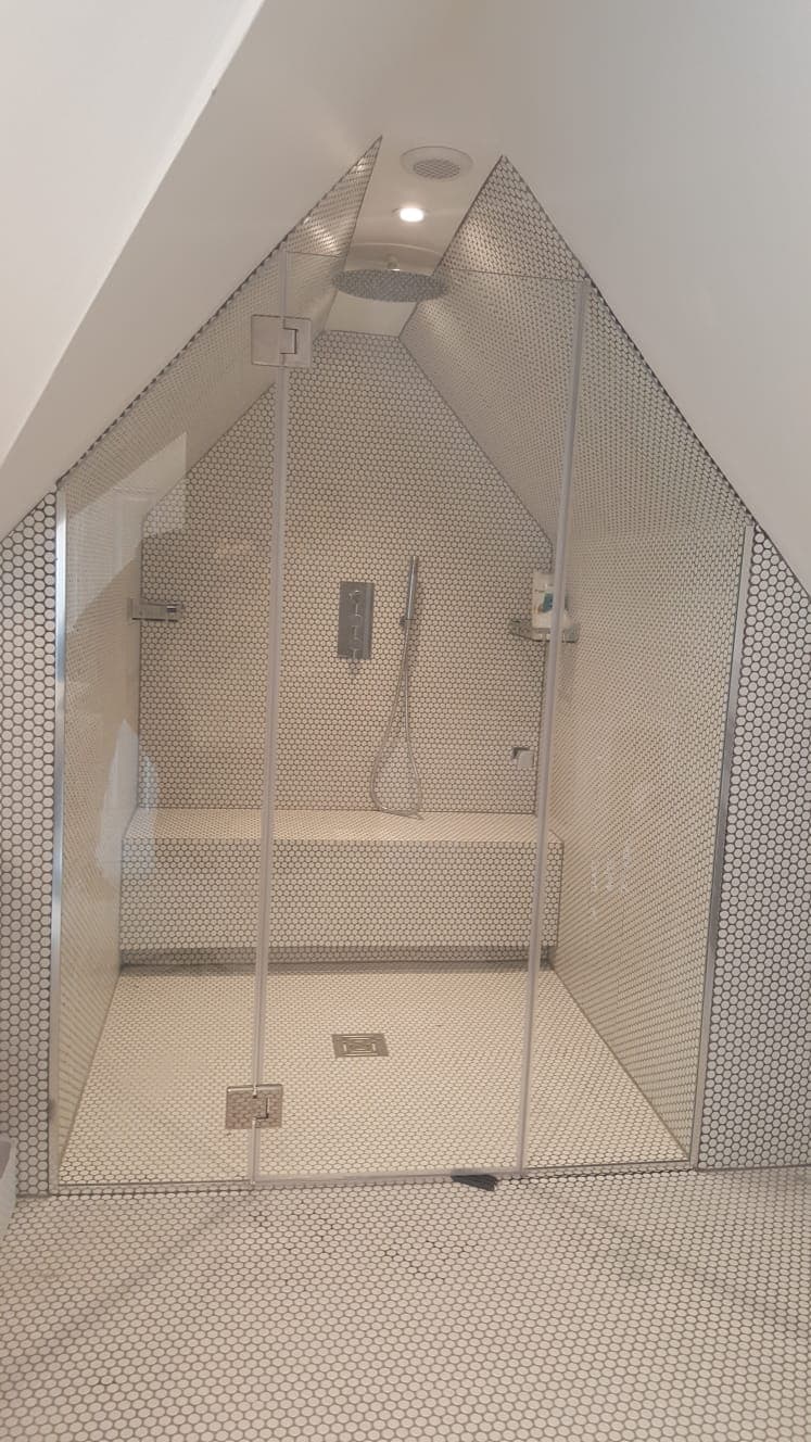 Shower enclosure under a sloping loft ceiling, the glass panel cut to follow the roof angle, with penny-round tiling