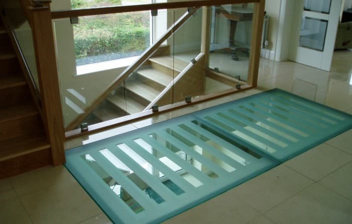 Interior walk-on glass floor panel with sandblasted anti-slip banding, beside a staircase with a glass balustrade and timber handrail